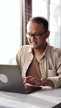Cheerful young Asian man is sitting down in a cafe while working using his laptop.