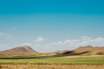 Obraz premium Panoramic view of the Iranian countryside featuring rolling green plains, golden grasslands, and rugged mountains beneath a vivid blue sky with scattered clouds.