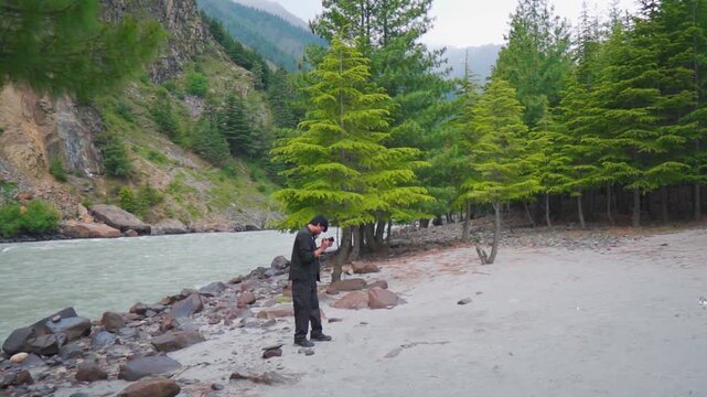 Wide angle shot of river flowing besides Pine tree forest with an Indian male travel influencer capturing beauty of nature with his camera while standing in the middle at Manali in Himachal, India.