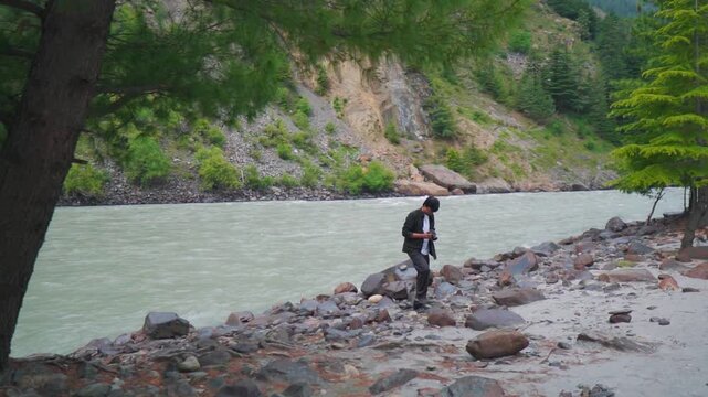 Wide angle shot of a male travel content creator taking videos with his camera besides flowing Chenab river and Pine trees at Mini Manali in Udaipur, Lahaul Valley, Himachal Pradesh, India.