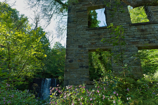 Stone ruins with open windows are framed by lush green trees and pink flowers, with a waterfall flowing nearby - Ashworth Valley, Rochdale, UK