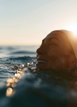 Close-up portrait of a woman's face in the ocean. she is wearing a white headscarf and is smiling widely, with her eyes closed and her mouth slightly open.
