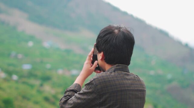 Rear view shot of an Indian male tourist taking videos of the mountains with his mobile phone during his trip to the Himalayas at Manali in Himachal Pradesh, India. Tourist enjoying his vacation. 