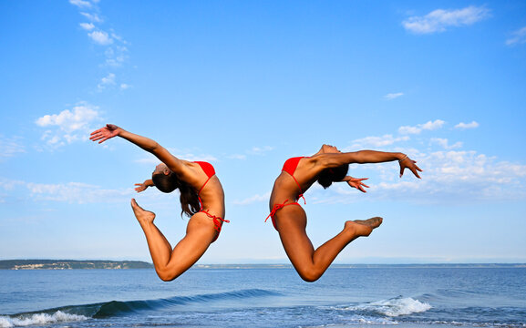 Two women perform synchronized partner yoga jumps on a beach