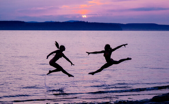 Two women perform acrobatic jumps in water at sunset on lake shore