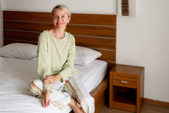 Woman sitting on bed in bedroom wearing pajamas. Happy caucasian female resting at home in morning. Lifestyle portrait of lady relaxing in cozy interior. Comfort and leisure concept.