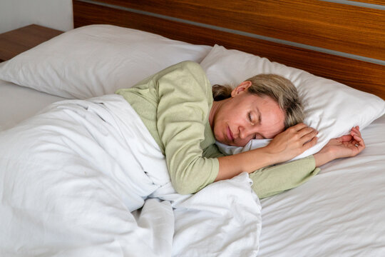 Woman sleeping in bed with head on soft white pillow. Young female resting under blanket in cozy bedroom. Calm nap time, healthy sleep and relaxation concept. Home morning routine.
