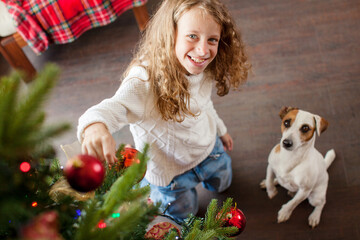 Obraz premium Happy child girl decorating Christmas tree with red ball ornament. Cute dog sitting nearby on floor. Winter holiday celebration at home with pet. Joyful childhood and family festive time.