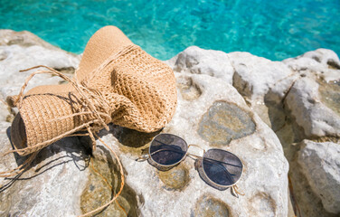 A straw beach bag and sunglasses resting on a rocky surface near clear turquoise water. Ideal for summer and vacation themes. © Mila