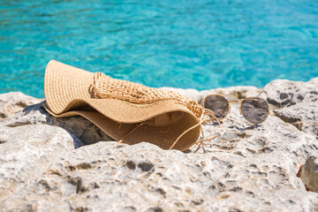A straw sun hat and sunglasses resting on a rocky surface by the clear blue water. Ideal for summer and beach themes. © Mila