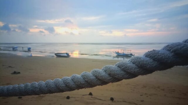 A thick nautical rope in close-up focus with a blurred tropical beach and fishing boats at sunset in the background.