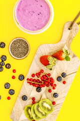 Fototapeta premium Preparing healthy fruit smoothie. Acai smoothie bowl near cutting board with fresh fruits, berries, chia seeds on yellow background top view