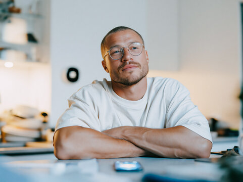 Thoughtful Young Man with Glasses Sitting with Arms Crossed in Bright Modern Office