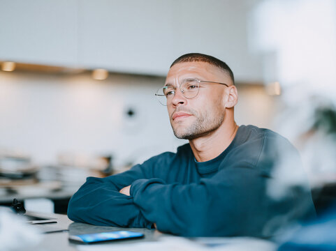 Thoughtful Young Man with Glasses Sitting with Arms Crossed in Bright Modern Office