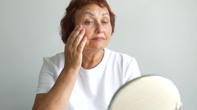 Portrait of elderly woman looking at herself in mirror while sitting at table in bright room. Adult woman examines wrinkles and touches face in cozy room. Concept of care, and enjoyment.