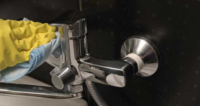 Cleaning the bathroom faucet, hands in yellow gloves wipe the faucet with a cleaning solution.