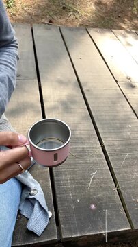 Slow motion close-up shot of woman's hands pouring a drink from a pink insulated thermos into a matching cup. Beautiful tall pine trees in the background. Perfect for camping or hiking themes.