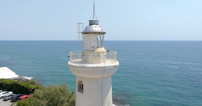 Close-up shot of the top lantern room and balcony of a white coastal lighthouse with a metal dome, set against a vast blue sea, clear sky, and green coastal vegetation on a sunny day.