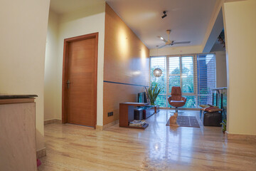Fototapeta premium Warm modern apartment interior featuring light marble flooring, a wooden accent wall, large floor-to-ceiling windows, a leather armchair, and a ginger cat sitting on a small rug.