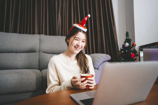 Young adult asian woman using laptop relax sitting in cozy winter living room with christmas tree enjoy alone at home for the holidays