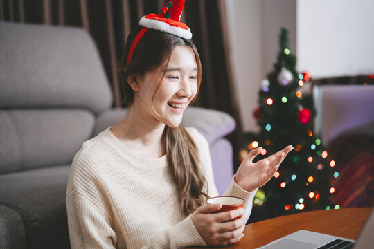 Young adult asian woman using laptop relax sitting in cozy winter living room with christmas tree enjoy alone at home for the holidays