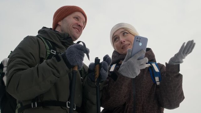 Caucasian couple hiking in winter, using smartphone for navigation and pointing at distance. Perfect for winter tourism, travel apps, and outdoor recreation marketing campaigns