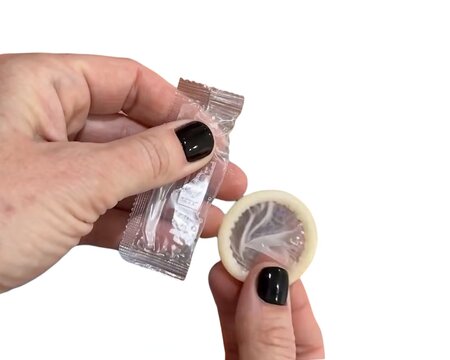 A person with black nail polish opening a silver condom wrapper and holding an unwrapped latex condom against a white background.