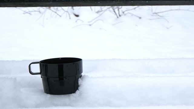 A black mug is placed on a snowy surface. A hand reaches towards the mug. The scene conveys a winter theme with cold weather elements.