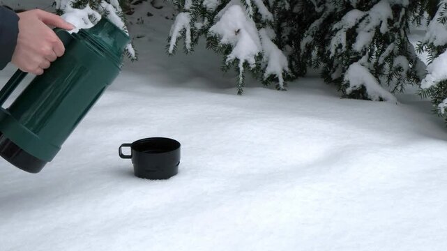 A black thermos mug is placed on a snowy surface. A gloved hand reaches for the mug. Snow-covered trees are in the background, creating a winter scene