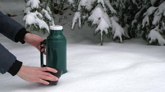 Green thermos and black mug on snowy ground as hands reach for the container, with snow-covered trees creating a cold outdoor winter scene.