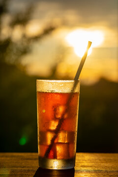Refreshing iced drink in tall glass with straw against warm sunset background, closeup. Ideal for summer vacation, beach bar menus, cafe advertising, and relaxation concepts