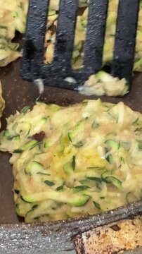 Zucchini fritters cooking in a pan, flipping golden vegetable pancakes with a spatula. Close-up overhead shot.