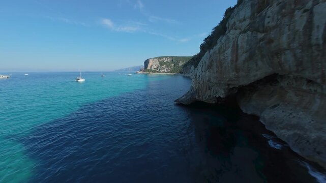 FPV drone flight low over turquoise water along Cala Luna's limestone cliffs, Baunei coast, Sardinia. Sea caves, crystal clear Mediterranean, dramatic coastline, June summer light.