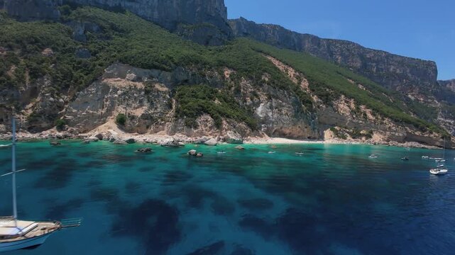 Aerial drone flight returning toward the dramatic cliffs of Cala Goloritze, Sardinia, passing a moored sailing yacht and a speeding boat in turquoise Mediterranean waters below.