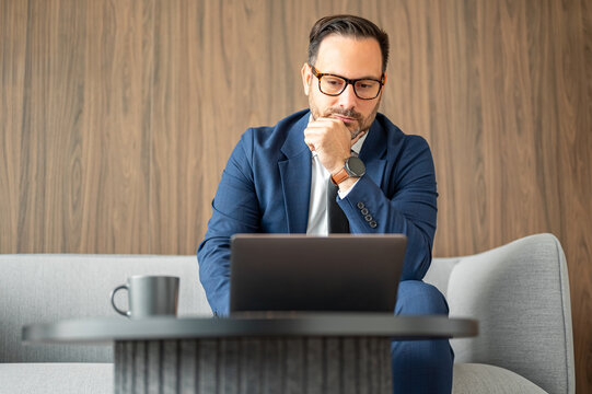 Professional adult man focusing on a laptop, sitting on a contemporary piece of furniture, contemplating business concepts in a corporate setting