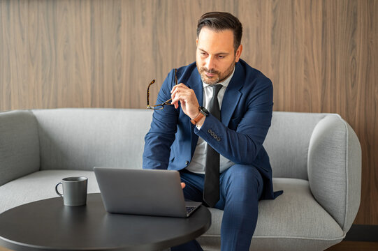 Businessman working, sitting on a modern upholstered bench, viewing content on a laptop with a hot beverage nearby, concentrating on online activities