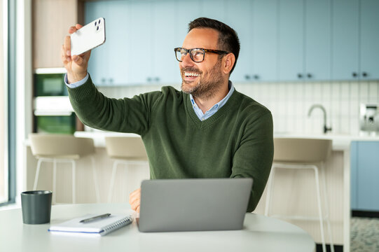 Smiling businessman using smartphone for a video call or social media while working remotely on a laptop, connecting from a modern kitchen setting