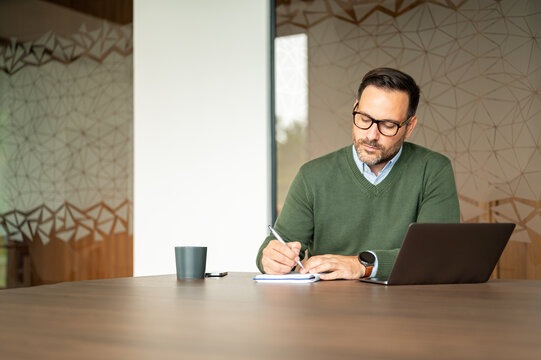 Professional adult man writing notes in a notebook at a dark table, using a laptop, a mug, and a mobile device in a contemporary workspace