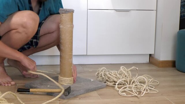 A man repairs a cat scratching post in a modern home. He is kneeling on the floor, using a hammer and rope. Tools and materials are scattered around.