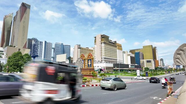 Timelapse of Phnom Penh traffic passing modern high-rises in Cambodia, with cars, scooters, roadside monument details and bright blue sky.