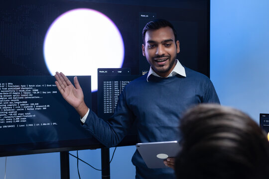 Software engineer holding laptop presents code displayed on large screen to colleague during collaborative programming session in office workspace focused on AI developer project review