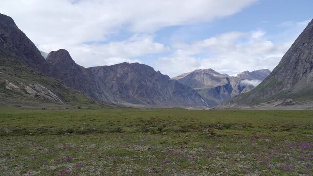 Wide symmetrical ground shot looking down a sweeping tundra valley floor toward jagged mountain peaks under a partly cloudy sky.