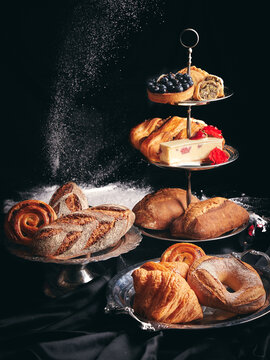 Set of French Artisanal Gourmet Bakery Brioche Bread Pastry Baked Goods Setup on Vintage Silver Cake Stand with Flour Throwing Behind Dark Background Setup on Black Cloth