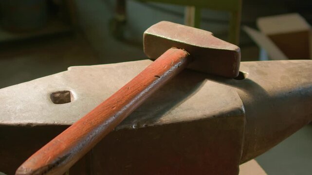 Blacksmith hammer anvil warm light beam illuminating workshop tools, handheld shot. Close up of traditional metalworking equipment on forge steel.