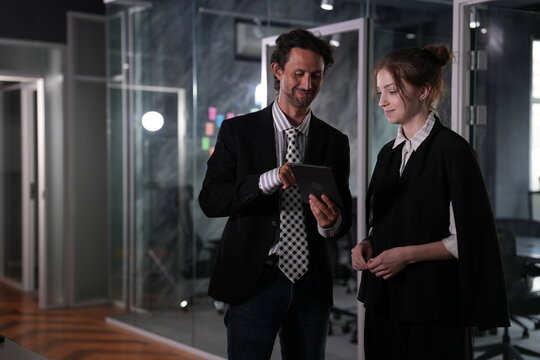 Grinning long haired mentor and softly smiling young woman review something amusing on the tablet amid dark glass cubicles and warm herringbone flooring in a bright modern coworking office.