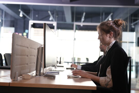 Young woman smiles faintly to herself while working her computer at the shared desk, warm skin tones glowing against the overexposed office windows in a bright modern coworking office.