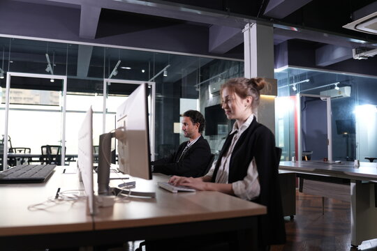 Both colleagues glance toward their screens in synchrony at the shared workstation, industrial ceiling and white window grids towering over the quiet scene in a bright modern coworking.