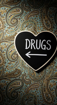 Black heart-shaped chalkboard with "DRUGS" and an arrow on a paisley patterned background, symbolizing direction or warning regarding substance use.