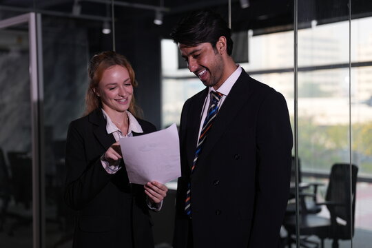 Businesswoman raises the document toward the window light so both colleagues can reread the key paragraph in the quiet dark toned office in a bright modern coworking office.