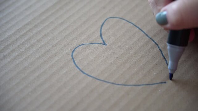 Hand drawing a heart with a blue highlighter on cardboard, representing love, warmth, or simple emotional expression, close-up